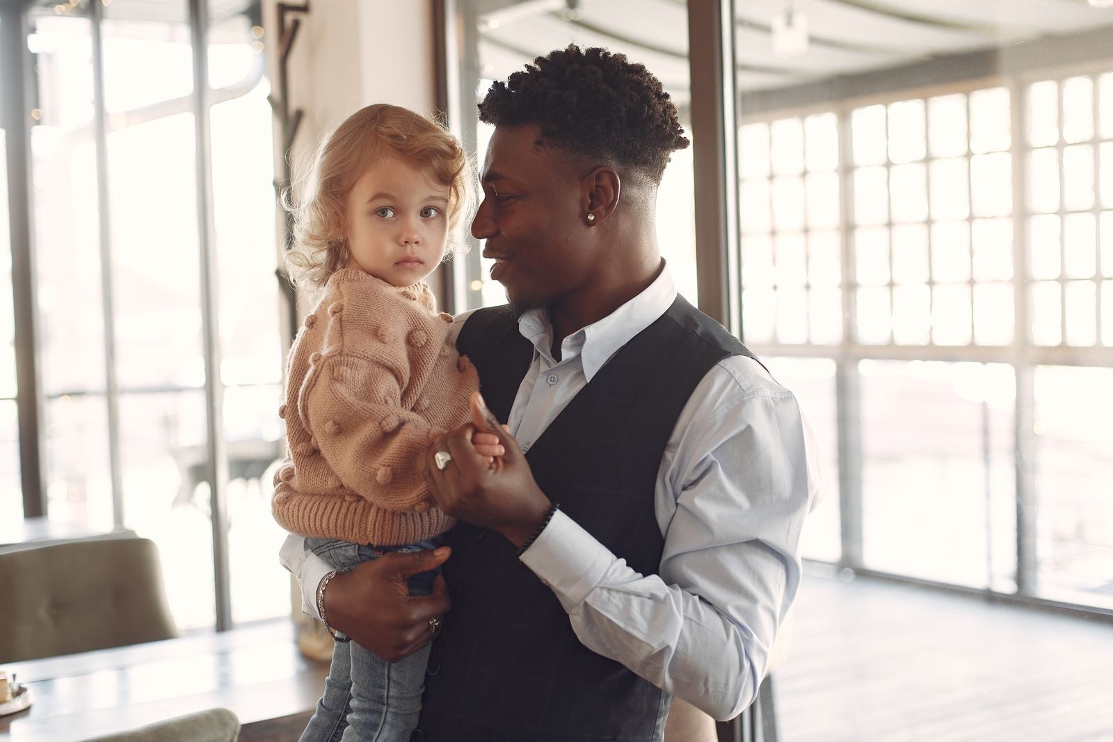 Positive African American man in formal suit holding cute little girl on hands while spending time together in modern light apartment